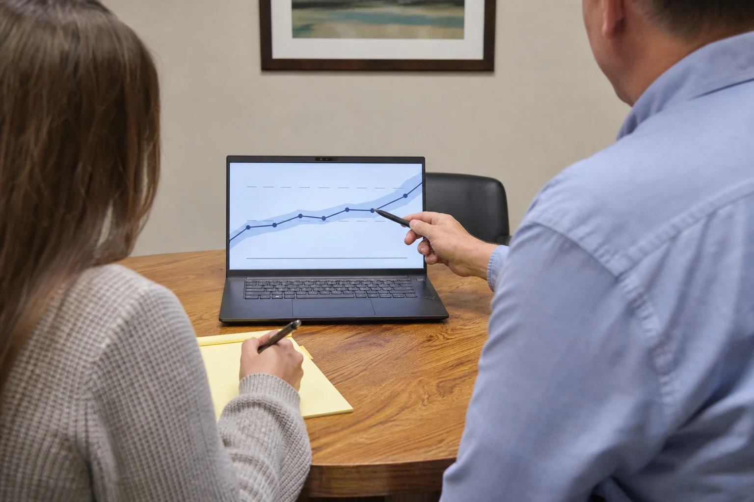 Agent in a blue shirt points a pen at a line chart on a laptop while a woman in a gray sweater takes notes on a yellow legal pad at a wooden table