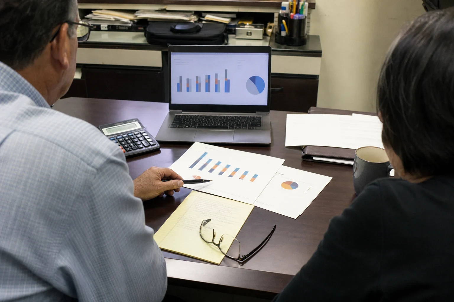 Two people seen from behind at a small office desk reviewing bar and pie charts on a laptop and printouts, with a calculator, legal pad, glasses and a mug